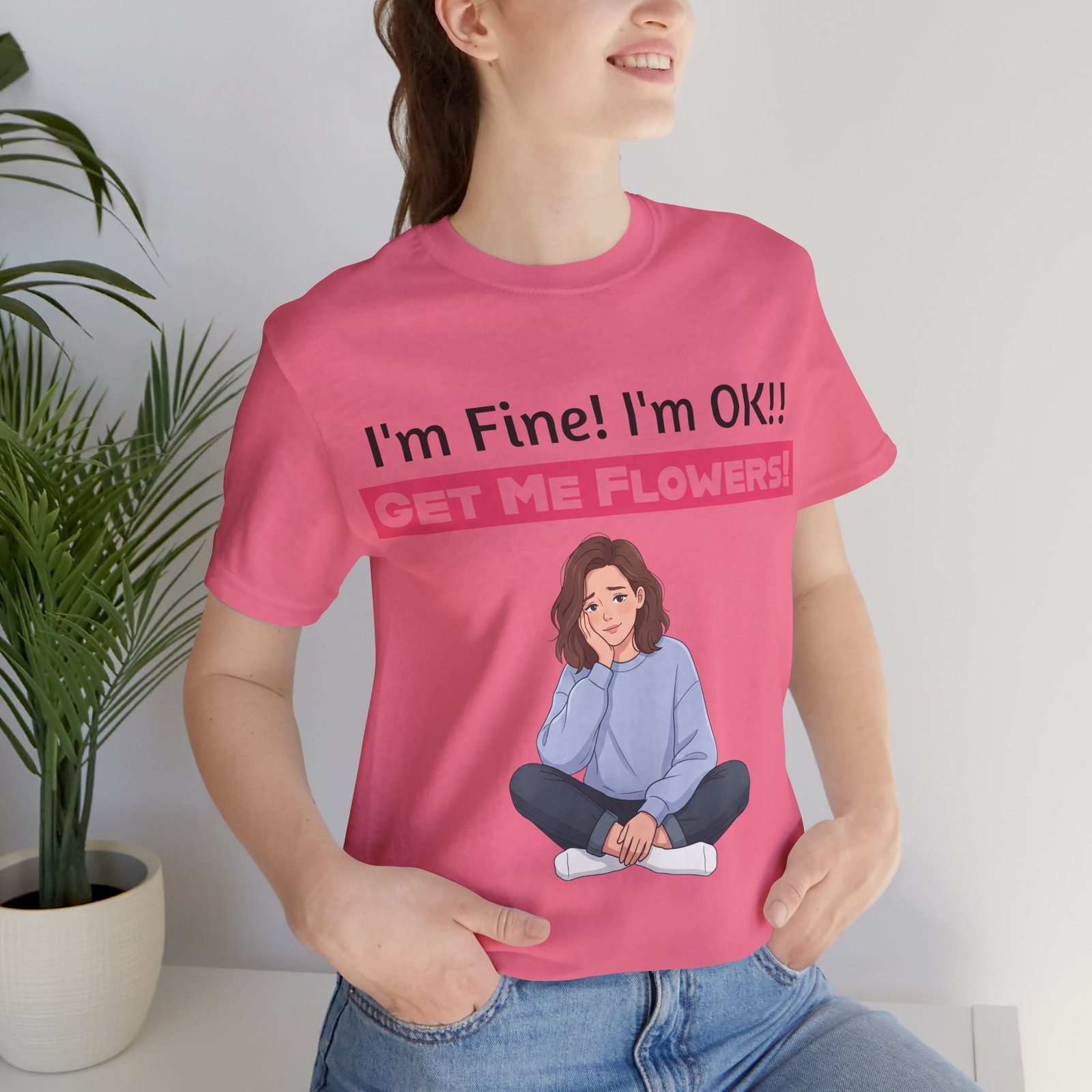 Woman wearing a pink funny T-shirt with 'I'm Fine! I'm OK!! GET ME FLOWERS!' text and a cartoon girl graphic.